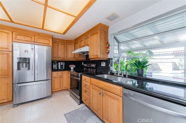 kitchen featuring a sink, under cabinet range hood, light tile patterned floors, stainless steel appliances, and dark stone counters