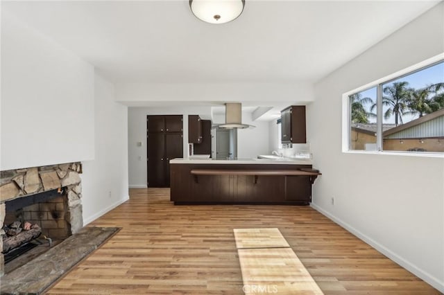 kitchen with light wood-style flooring, dark brown cabinets, a fireplace, baseboards, and a peninsula