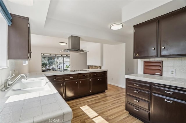 kitchen featuring wall chimney range hood, a sink, tile counters, dark brown cabinets, and light wood finished floors
