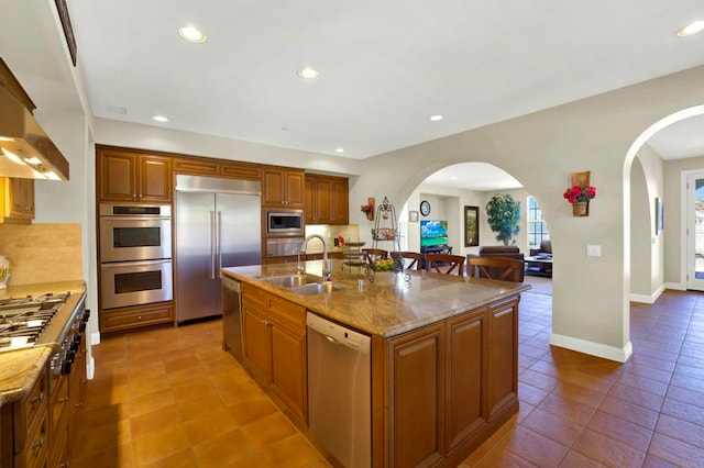 kitchen with light stone counters, a sink, built in appliances, arched walkways, and tile patterned flooring