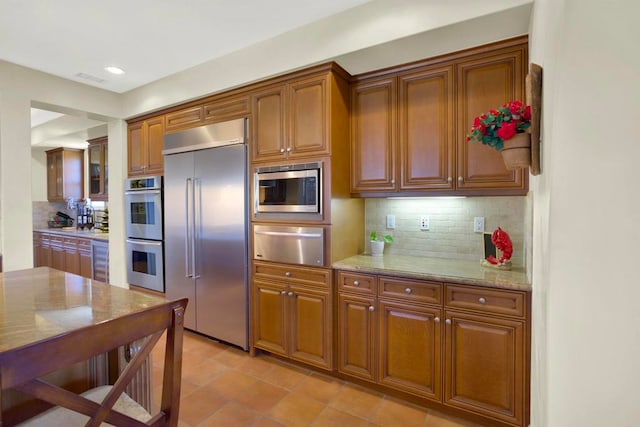 kitchen featuring light stone counters, brown cabinetry, a warming drawer, tasteful backsplash, and built in appliances