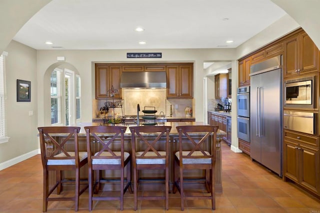 kitchen featuring arched walkways, a warming drawer, backsplash, built in appliances, and under cabinet range hood