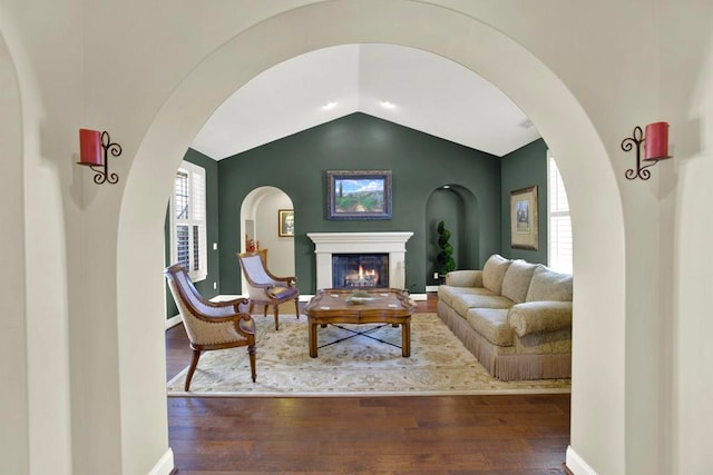 living room featuring arched walkways, vaulted ceiling, wood finished floors, and a lit fireplace