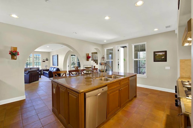 kitchen with arched walkways, a sink, brown cabinets, an island with sink, and appliances with stainless steel finishes