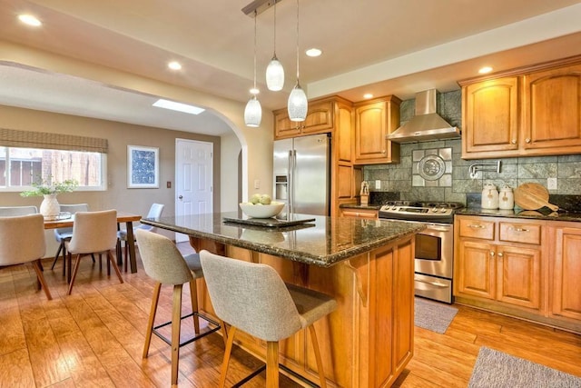 kitchen featuring light wood-type flooring, a breakfast bar, wall chimney exhaust hood, stainless steel appliances, and arched walkways