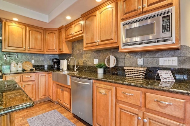 kitchen with light wood-style floors, dark stone counters, appliances with stainless steel finishes, a sink, and backsplash