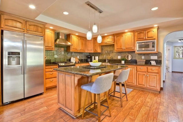 kitchen featuring light wood-type flooring, wall chimney exhaust hood, a raised ceiling, stainless steel appliances, and arched walkways