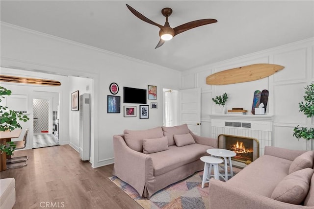 living area with a ceiling fan, wood finished floors, a brick fireplace, baseboards, and ornamental molding