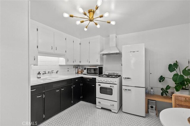 kitchen featuring white appliances, an inviting chandelier, dark cabinetry, a sink, and wall chimney exhaust hood