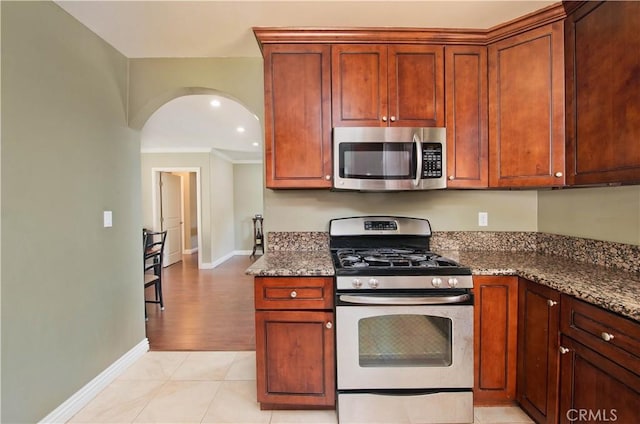 kitchen featuring light tile patterned floors, stainless steel appliances, baseboards, dark stone counters, and arched walkways