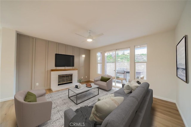 living area featuring a glass covered fireplace, light wood-type flooring, baseboards, and ceiling fan