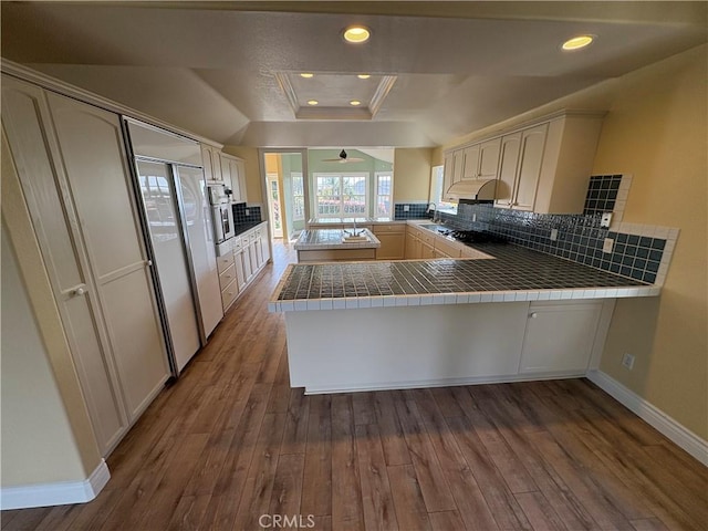 kitchen with dark wood finished floors, under cabinet range hood, a tray ceiling, baseboards, and a peninsula