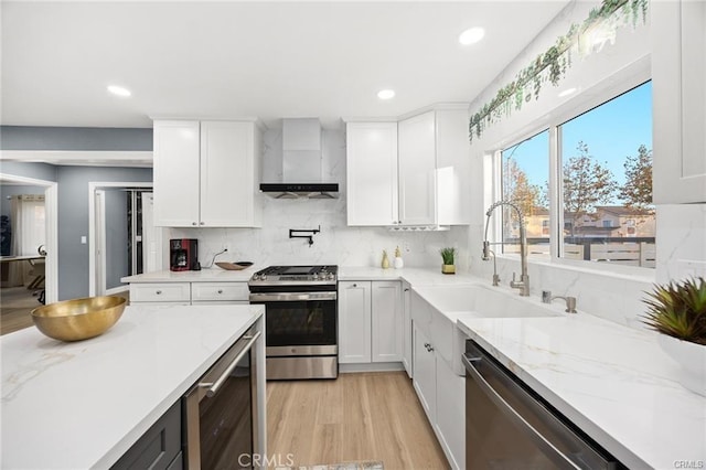 kitchen featuring white cabinets, a sink, light wood-type flooring, wall chimney exhaust hood, and appliances with stainless steel finishes