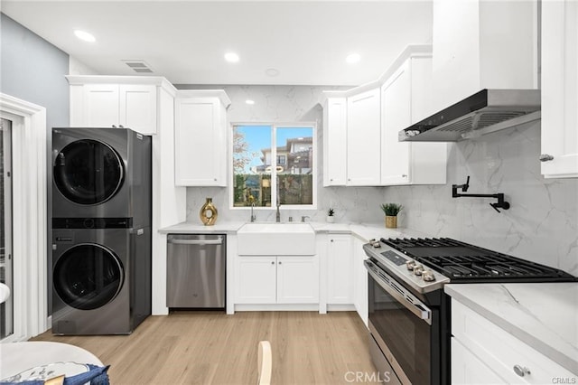 kitchen featuring wall chimney exhaust hood, stainless steel appliances, stacked washer and dryer, a sink, and white cabinetry