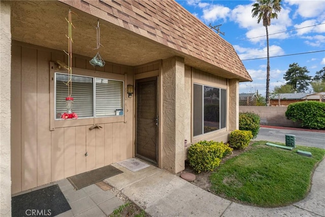entrance to property featuring stucco siding, fence, mansard roof, and roof with shingles