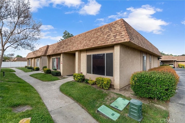 exterior space with stucco siding, mansard roof, fence, a shingled roof, and a lawn