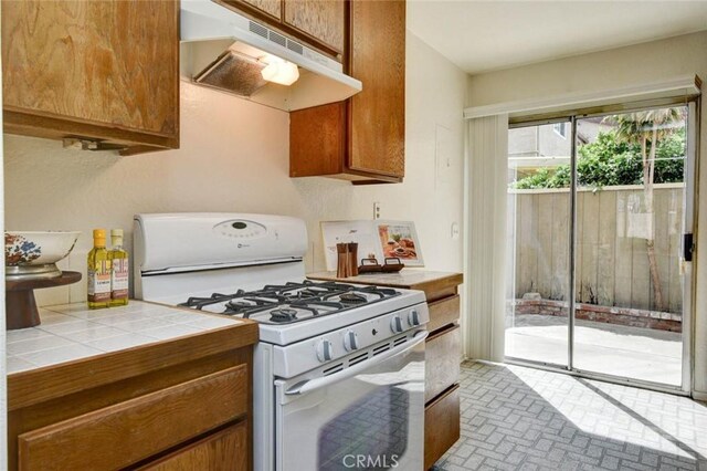kitchen with tile countertops, white gas range, brick patterned floor, under cabinet range hood, and brown cabinets