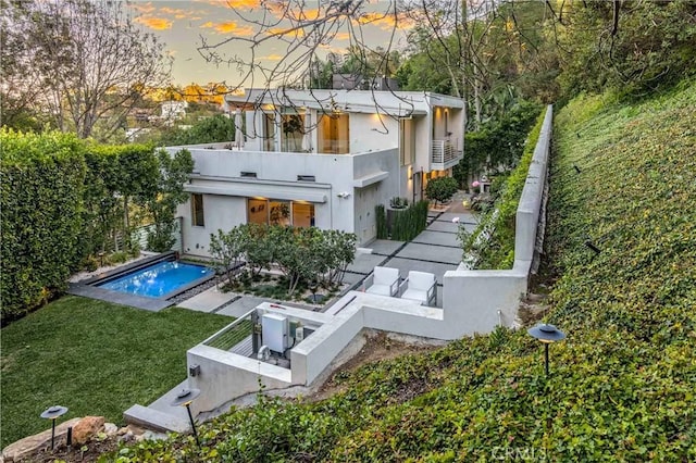 back of house at dusk featuring an outdoor pool, a balcony, fence, and a lawn