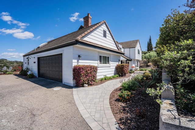 view of property exterior with a tile roof, a chimney, driveway, an attached garage, and fence