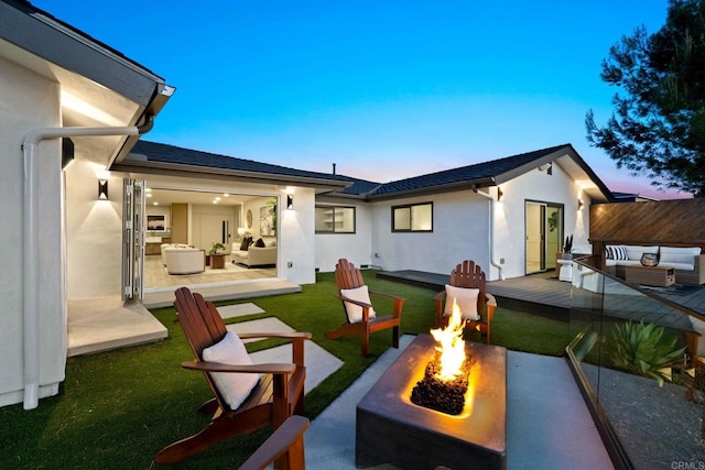back of property at dusk with stucco siding, an outdoor living space with a fire pit, a lawn, and a wooden deck
