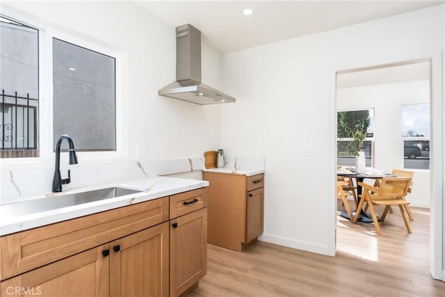 kitchen featuring a sink, light stone countertops, wall chimney range hood, baseboards, and light wood-style floors