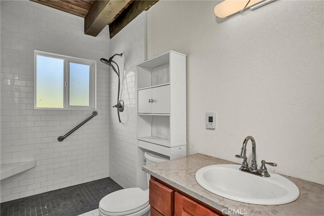 full bathroom with a tile shower, toilet, beam ceiling, vanity, and a textured wall