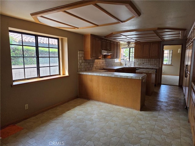 kitchen with a sink, tile countertops, a peninsula, decorative backsplash, and under cabinet range hood