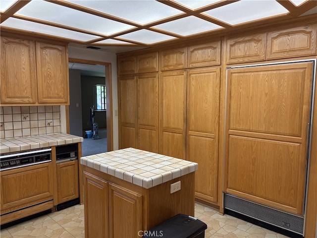 kitchen with tile counters, a center island, decorative backsplash, paneled dishwasher, and visible vents