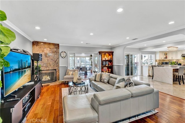 living room with crown molding, recessed lighting, arched walkways, light wood-style flooring, and a stone fireplace