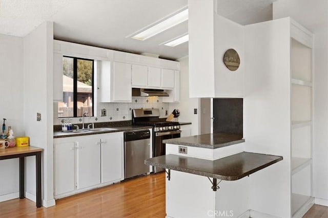kitchen with a sink, appliances with stainless steel finishes, dark countertops, light wood-style floors, and under cabinet range hood