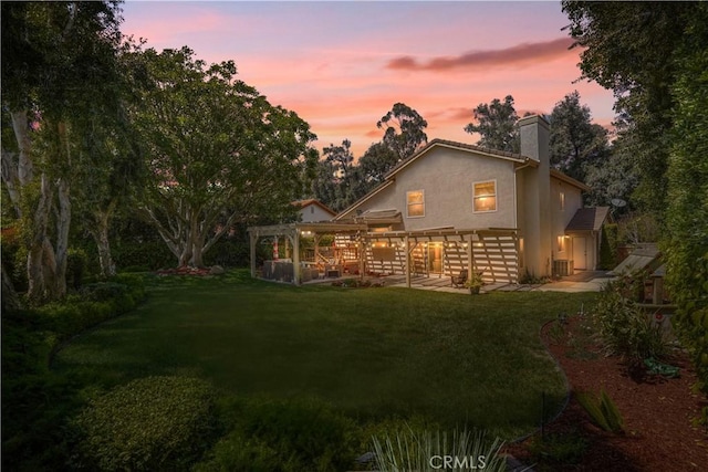 back of house at dusk with a yard, central AC unit, stucco siding, and a chimney