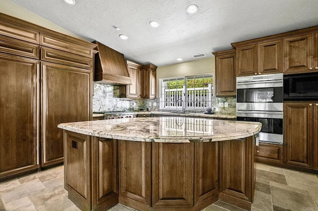 kitchen featuring stone tile flooring, stainless steel double oven, custom exhaust hood, and backsplash