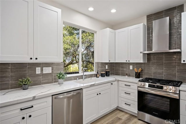 kitchen featuring white cabinets, wall chimney exhaust hood, a sink, light stone countertops, and stainless steel appliances