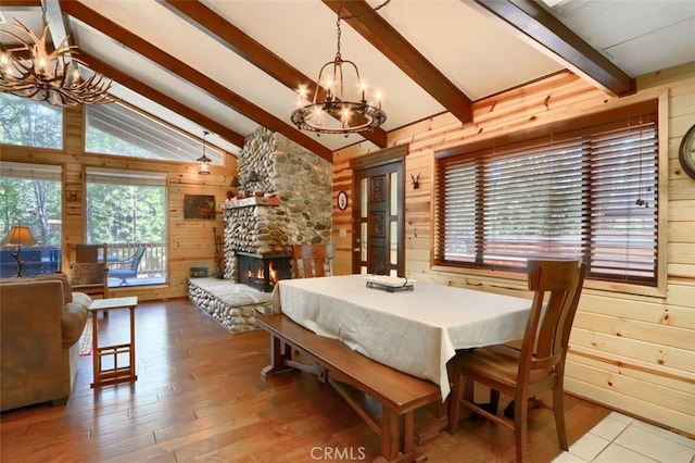 dining space featuring vaulted ceiling with beams, wood finished floors, wood walls, a stone fireplace, and an inviting chandelier