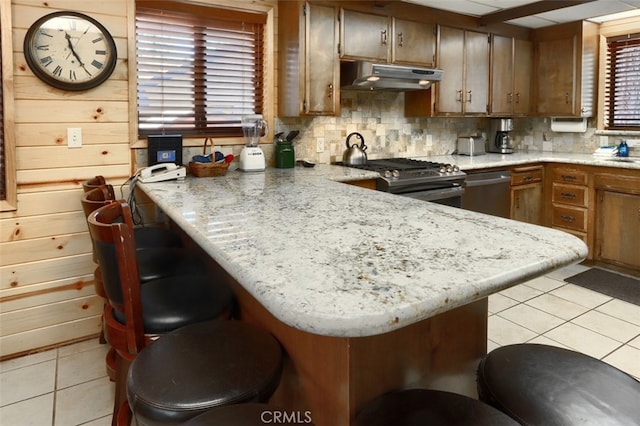 kitchen featuring light tile patterned flooring, tasteful backsplash, appliances with stainless steel finishes, a peninsula, and under cabinet range hood