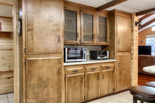 kitchen with stainless steel microwave, light countertops, brown cabinets, glass insert cabinets, and light tile patterned floors