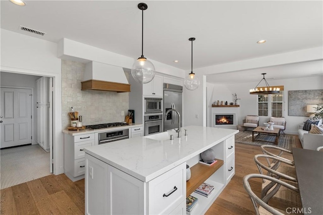 kitchen featuring visible vents, built in appliances, custom range hood, light wood-style flooring, and a glass covered fireplace