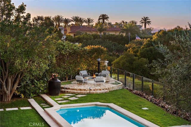 pool at dusk with a patio, a fire pit, an outdoor pool, fence, and a lawn
