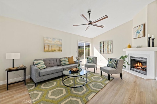 living room featuring lofted ceiling, a lit fireplace, baseboards, light wood finished floors, and a ceiling fan