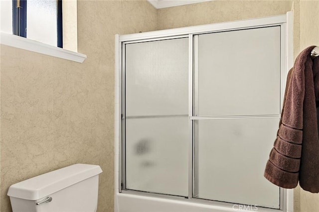 bathroom featuring a textured wall, combined bath / shower with glass door, and toilet