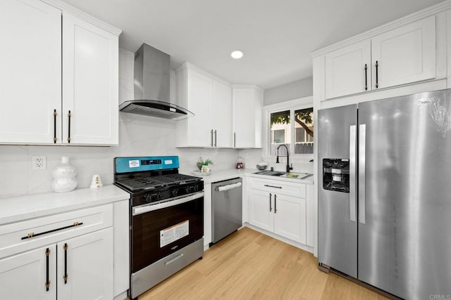 kitchen featuring a sink, light wood finished floors, appliances with stainless steel finishes, wall chimney range hood, and white cabinets