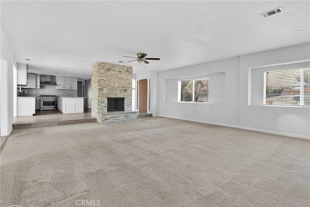 unfurnished living room featuring light carpet, a stone fireplace, baseboards, visible vents, and ceiling fan