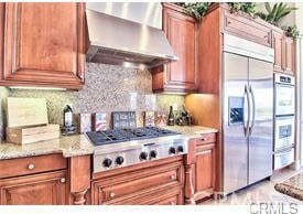 4069 Nancy Cir, Corona, CA 92881 - kitchen featuring wall chimney exhaust hood, stainless steel appliances, brown cabinets, decorative backsplash, and light stone countertops