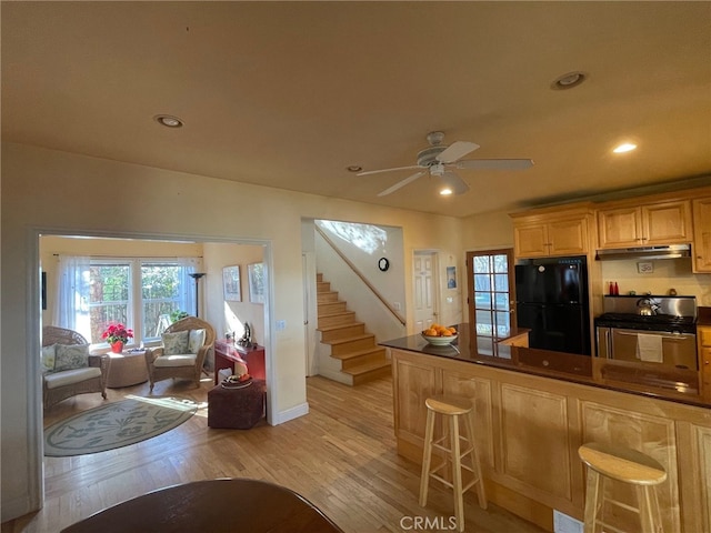 kitchen with freestanding refrigerator, under cabinet range hood, dark countertops, gas range oven, and light wood-type flooring