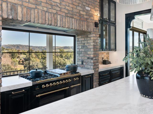 2255 Via Dulcea, Fallbrook, CA 92028 - kitchen featuring stainless steel stove, dark cabinetry, glass insert cabinets, plenty of natural light, and brick wall