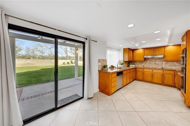 kitchen featuring a sink, brown cabinetry, a raised ceiling, under cabinet range hood, and tasteful backsplash