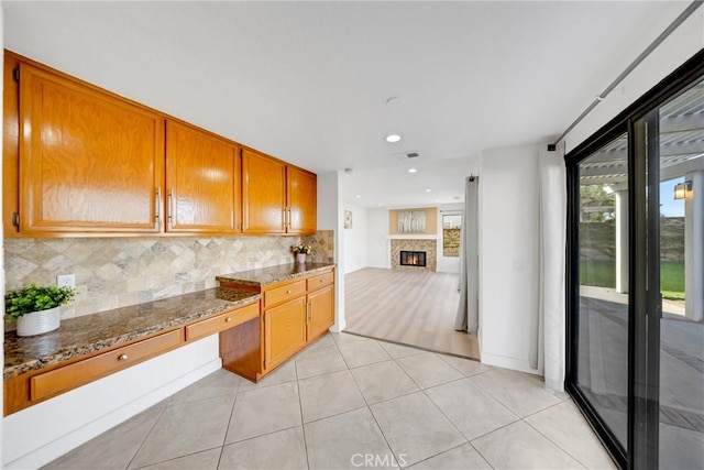 kitchen featuring brown cabinetry, dark stone countertops, recessed lighting, decorative backsplash, and a glass covered fireplace