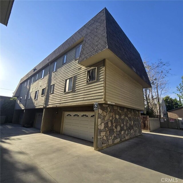 view of side of property featuring driveway, a garage, fence, a shingled roof, and stone siding