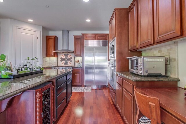 kitchen featuring wall chimney range hood, tasteful backsplash, built in appliances, dark wood-style floors, and recessed lighting