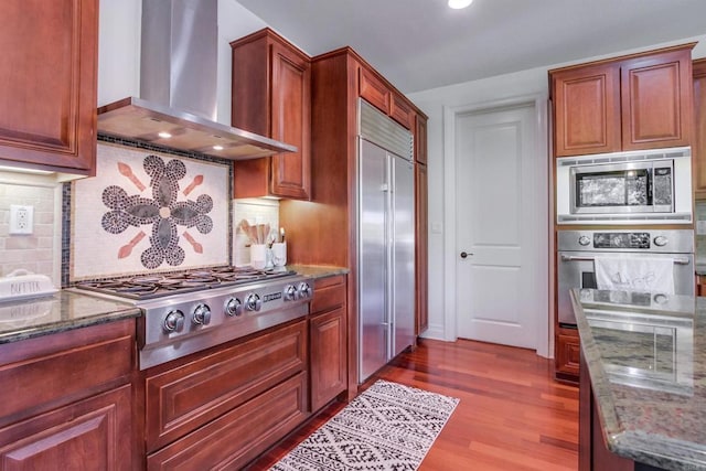 kitchen featuring built in appliances, wood finished floors, dark stone countertops, and wall chimney range hood